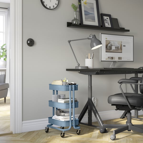 Blue RÅSKOG trolley with 3 shelves next to desk. Computer, lamp, chair visible.