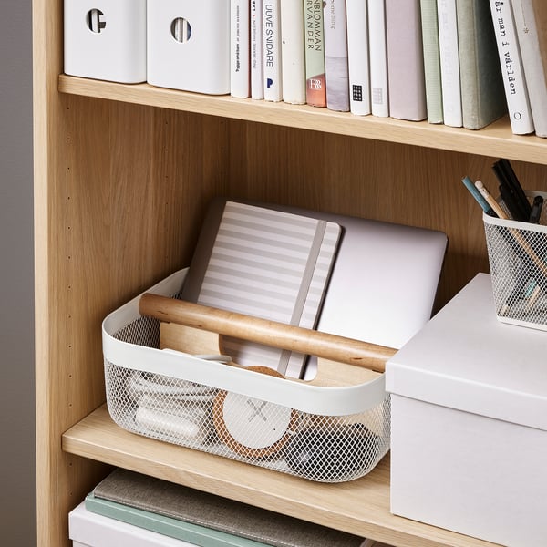 Wooden bookshelf with RISATORP storage basket holding a laptop, notebook, and pens, ideal for organising and transporting small items.