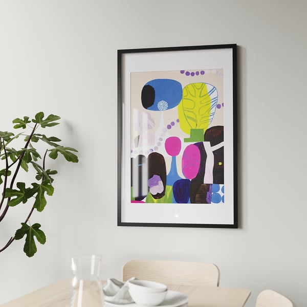 The framed artwork, RÖDALM, hang on a wall. Its bright, abstract design fits in a black frame, bigger than the chair beneath it and nearby plant.