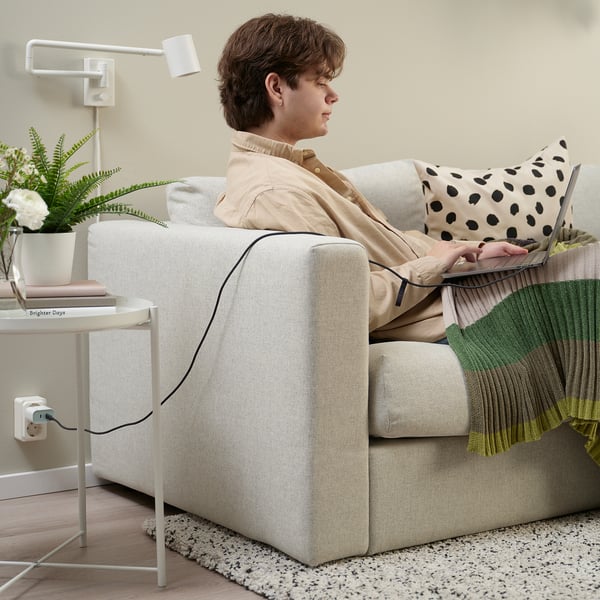 A person sits on a beige sofa, using a laptop with a usb-c cable connected. A side table with a lamp, books, and a plant is nearby.