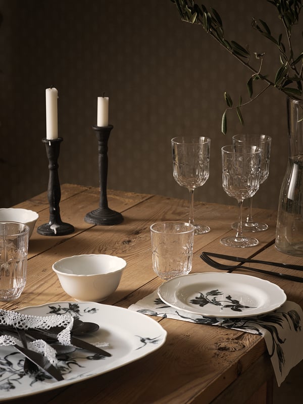 Timeless table setting with SÄLLSKAPLIG glassware, white plates, black cutlery, black candlesticks, and vine branch.