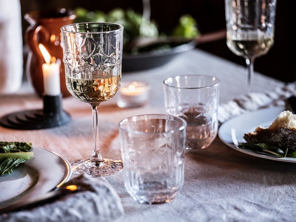 Elegant table setting with SÄLLSKAPLIG glassware, including wine glasses and tumblers, placed on a tablecloth beside plates of food and lit candles.