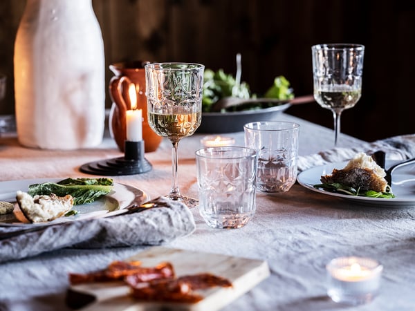 Table set with SÄLLSKAPLIG glassware, plates, and candles. Elegant crystal glasses, plates with food, grey cloth, white napkins, and warm candlelight.