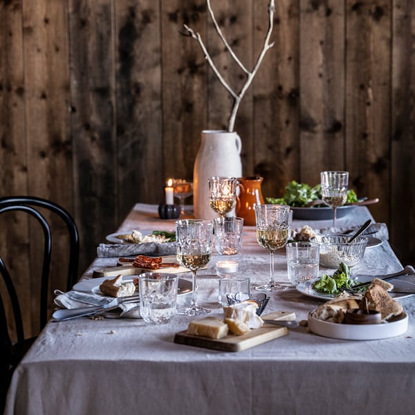 Elegant table set with SÄLLSKAPLIG clear glasses, candles, cheese, bread, on white linen. Rustic wooden backdrop.