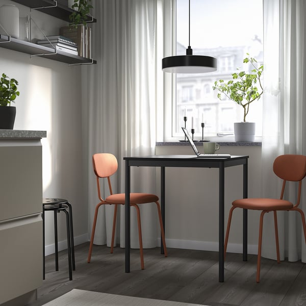 Minimalist workspace: black sandberg table, orange chairs. Metal base, upholstered seats. Bright, airy, with plants.