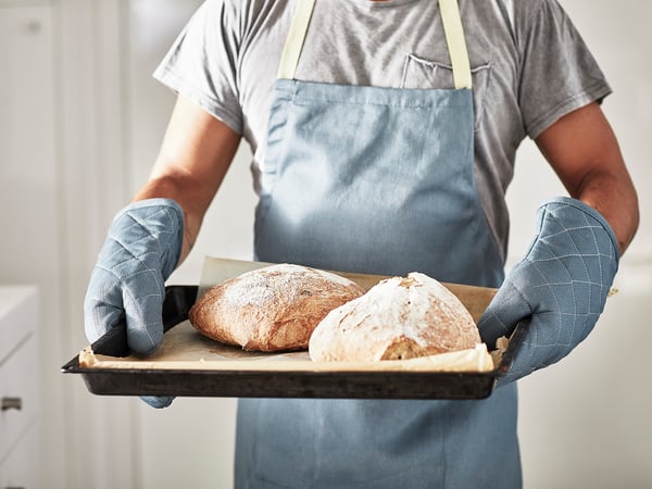 A person holds two loaves of bread on a tray, wearing a grey SANDVIVA apron and blue oven mitts.