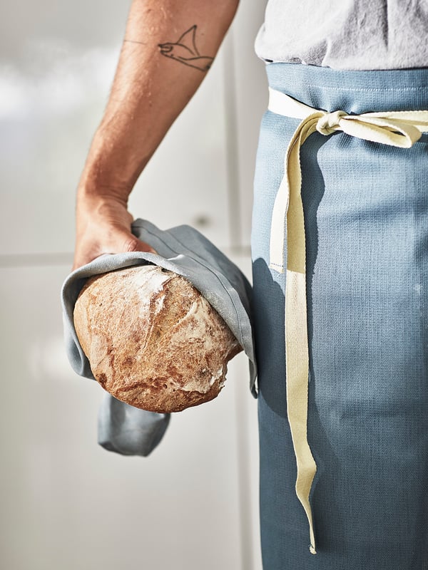 Person holding bread with tattooed arm, wearing blue and white apron with long ties.
