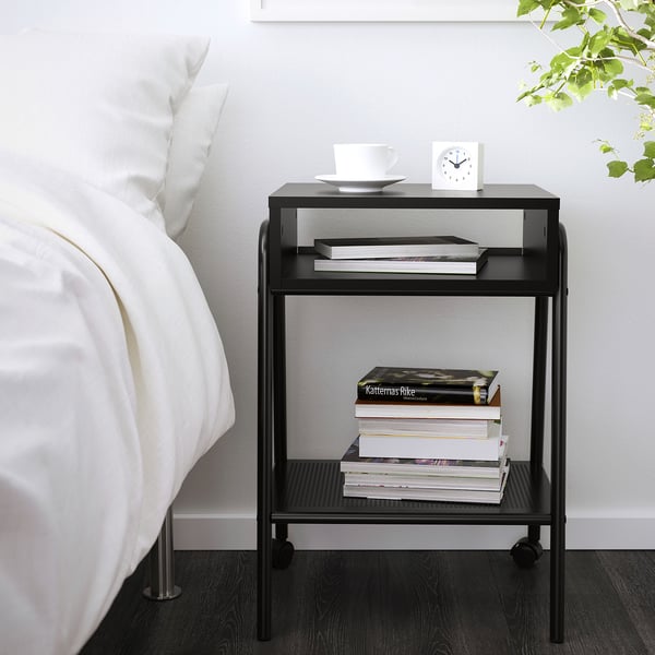 Small black metal bedside table beside a bed with books, white bowl, and alarm clock.