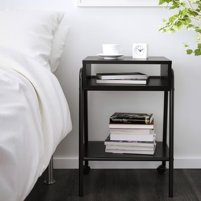Small black metal bedside table beside a bed with books, white bowl, and alarm clock.