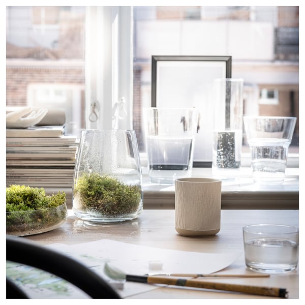 Desk near window with books, glass vases, SILKESTRÄD beige candle, green plant, notebook, pencil, black pen. Clean, organised workspace.