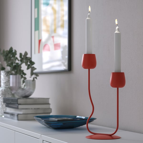 Red candlestick with two lit candles on a shelf, next to a plant and books.