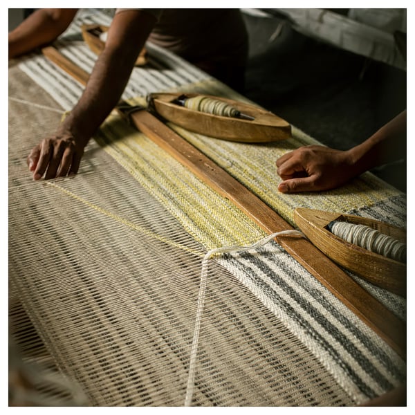 Close-up of hands weaving yellow and grey striped wool rug on loom, tweezers and threads visible.