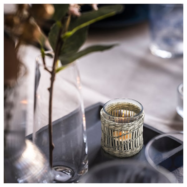 A SLIPPRIG tealight holder made of woven sedge with a lit candle, placed next to a clear glass vase with greenery on a dark table.