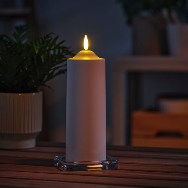 Single tall, white SMÅLOM led candle on wooden table, lit, surrounded by blurry plant and interior.