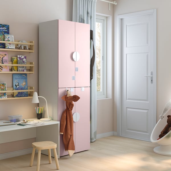 A kids room with a pink SMÅSTAD wardrobe has multiple shelves with colourful books, a desk with a lamp, and a small chair.