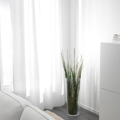 A minimalist living room with a white sofa; a vase of dried twigs stands near sheer white curtains, allowing natural light to philtre through.