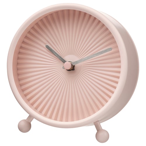 A pink, round SNOFSA clock with a sunburst design and silent quartz movement. It has a glass front, steel frame, and three spherical feet.