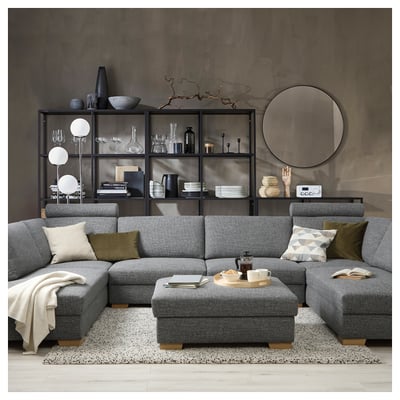Modern grey sectional sofa against dark wall, filled with cushions; displayed with coffee table, rug, and decorated shelves.