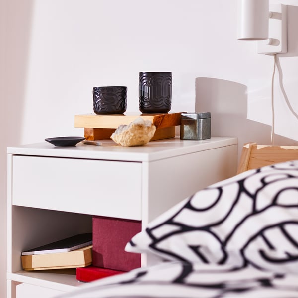 Black SÖTRÖNN candle in a small, dark stoneware jar sits on a white bedside table, smaller than a stack of books, near a patterned pillow.