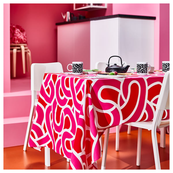 Bright pink tablecloth with white swirls on a small dining table with white chairs, set with cups and teapot.