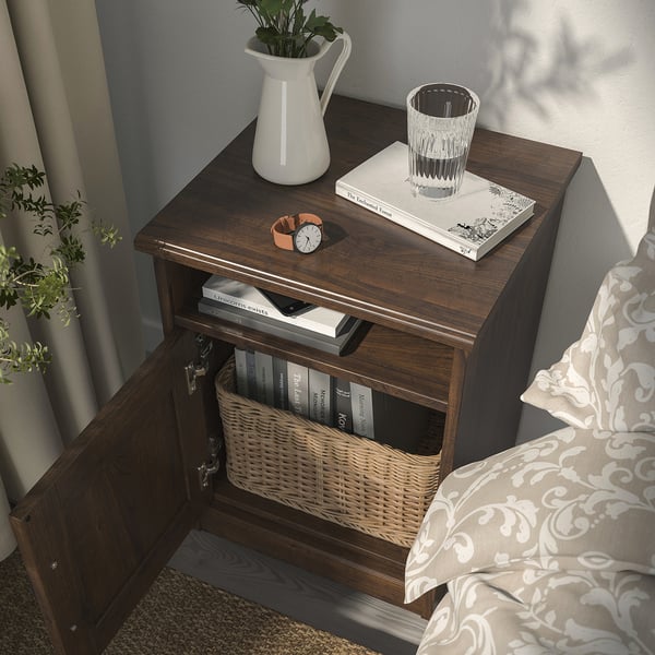 Wooden bedside table with drawer open, basket, books, vase, and clock, near bed with stylish fabric design.
