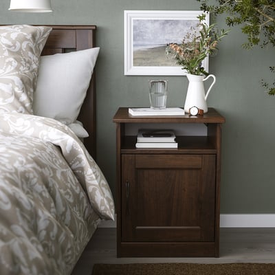 Small dark wooden bedside cabinet beside bed. Top holds books, vases, flowers, and a picture frame.