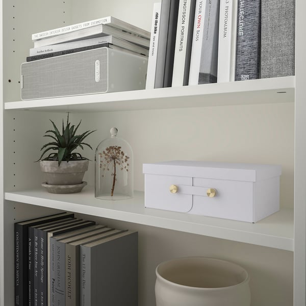Bookshelf with books, a speaker, potted plant, and a white storage box with gold handles.