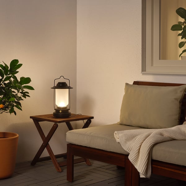 Small black outdoor lamp sits on small table, beside houseplant and bench with pillows.