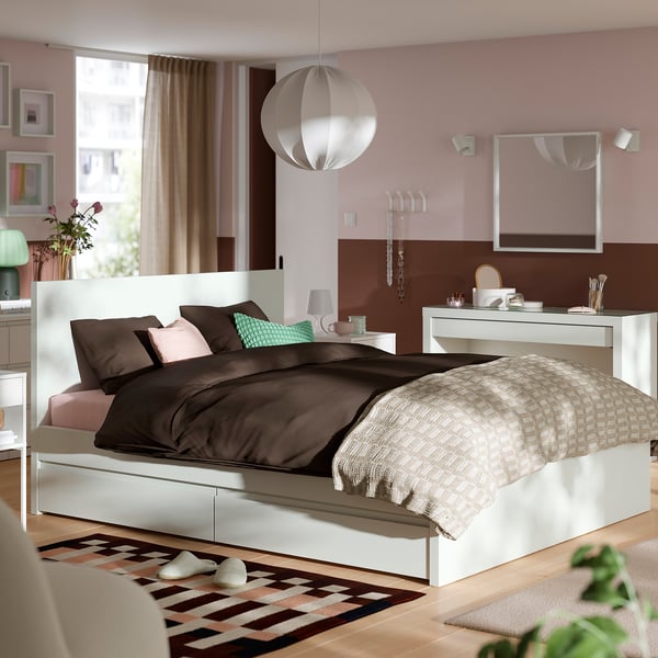 Modern bedroom: white bed, dark bedding, pastel pillows, pink walls, lamp, window, rug, art.