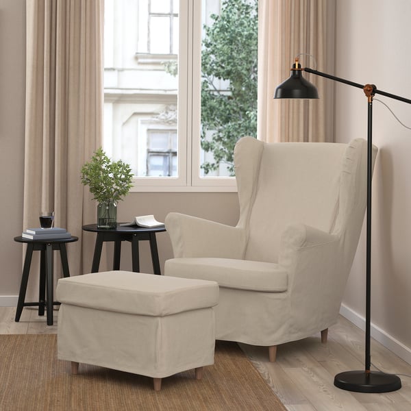 A cosy corner with a beige STRANDMON wing chair, matching footstool, black floor lamp, and side table with greenery and books, exuding warmth.