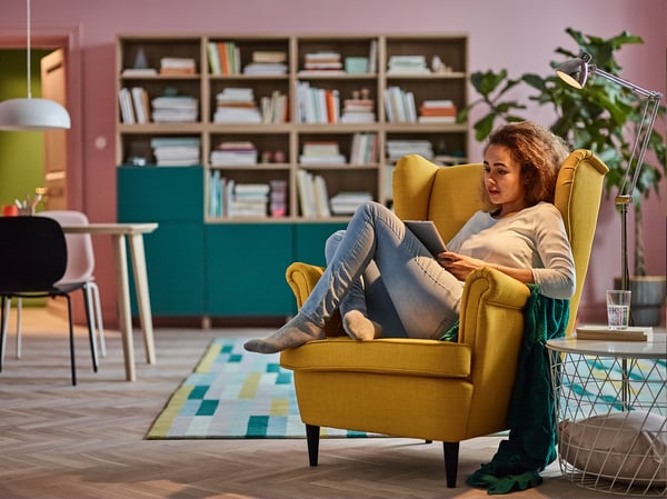 Person relaxing in yellow STRANDMON armchair, reading, surrounded by bookshelves and plants.