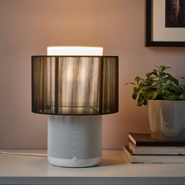Modern wifi lamp with woven base & striped shade, playing music next to books & a plant.