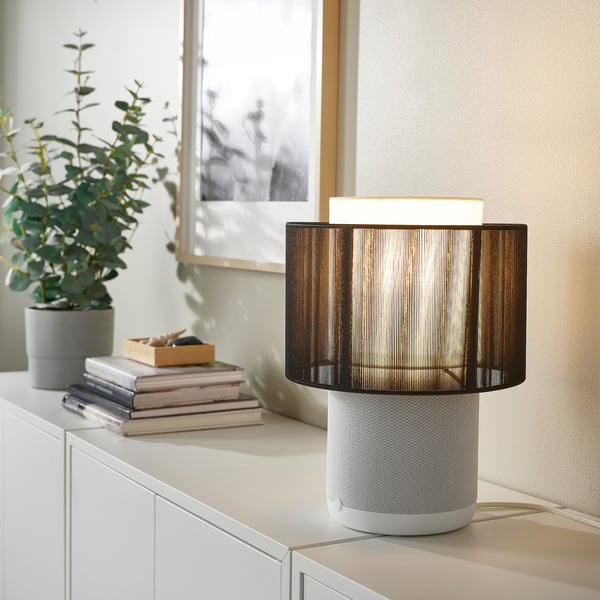 SYMFONISK white speaker lamp blend on shelf, surrounded by small books and potted plant, emitting soft light.