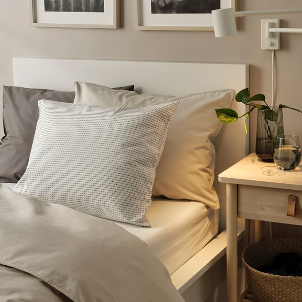 Cosy bed with white, grey, & tan pillows. Bedside table with water & plant in earthy tones.