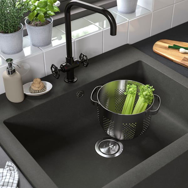 Black farmhouse sink with a strainer full of celery and parsley, featuring a modern matte black tap and cleaning supplies.