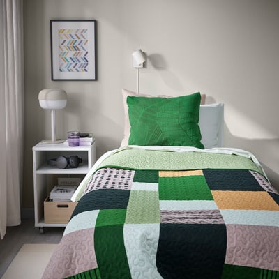 A bed with a TALLSVÄRMARE green patchwork bedspread, beside a small table with a lamp, books, and a picture above.