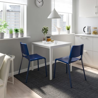 Cosy dining area with 2 blue TEODORES chairs, matching white table, by window with plants and a clock.