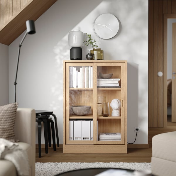 Wooden TONSTAD unit in light oak with sliding glass doors, shelves holding books, bowls, and vases, white clock, and decor atop.