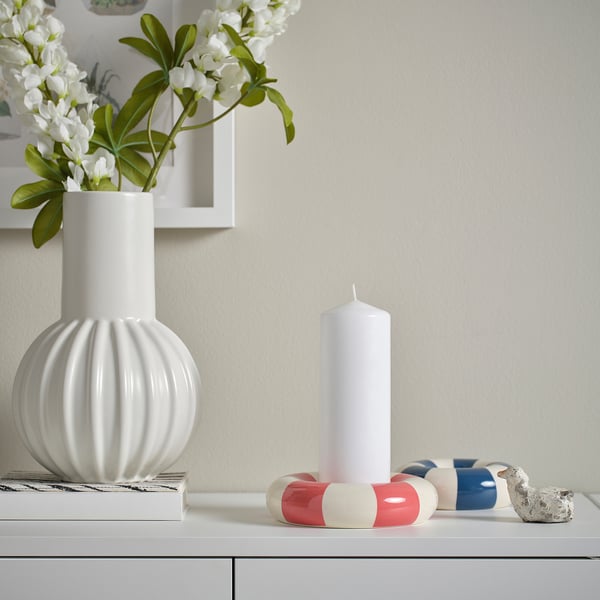 A TRÄDSTAM white candle holder like a swim ring sits small on a shelf, accompanied by a larger white candle and decorative objects.