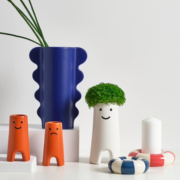 Colourful ceramic decor: blue vase, orange figures, white candle, green plant, white pillars. Happy, sad faces; swim ring design.