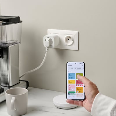 Hand holding smartphone with smart home app controlling plugged appliance via TRETAKT, coffee maker, and mug on counter.