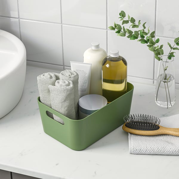 Green storage box on counter, holding towels, bottles, and cup. Organises bathroom items neatly.