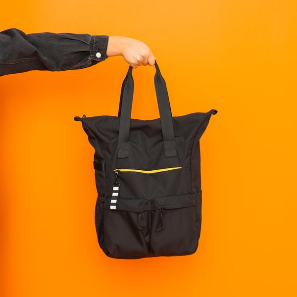 Hand holds black VÄRLDENS backpack, bright orange background. Has yellow accents, multiple pockets, made of recycled polyester.