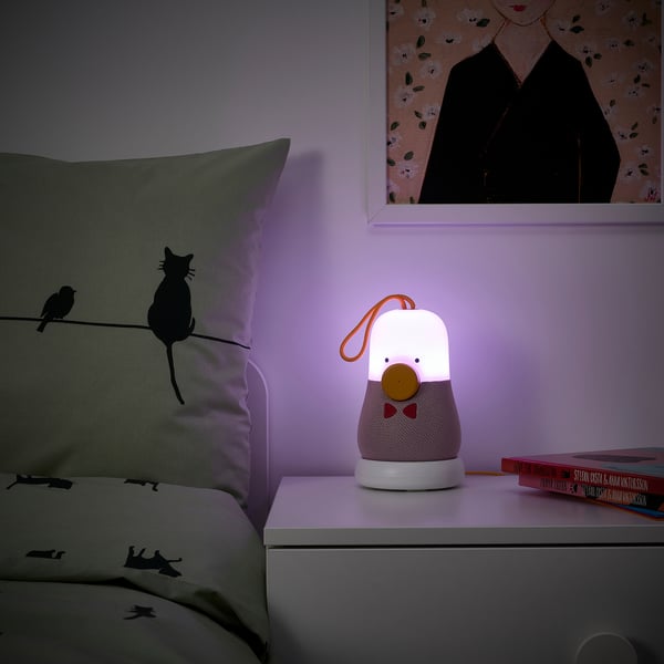 Kids lamp speaker combo emits soft purple light. Friendly character design on bedside table with pillow and book.