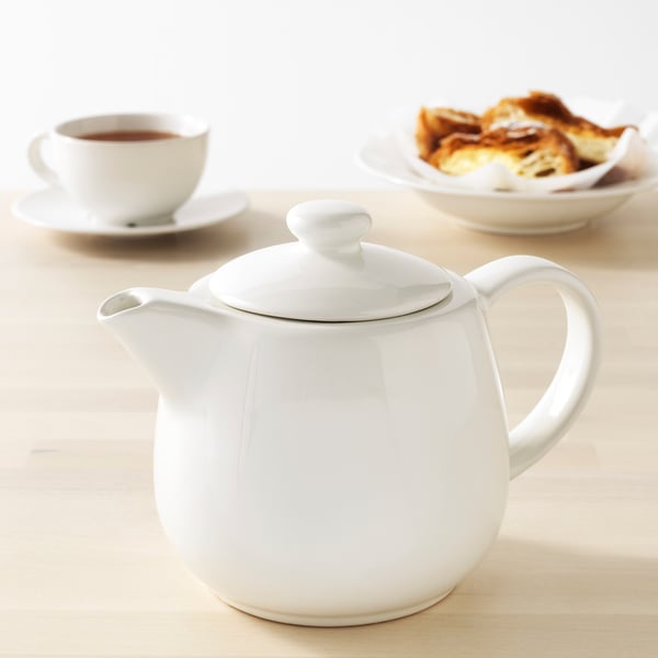 A white ceramic teapot with a lid and handle on a wooden table accompanied by a cup of tea and pastries.