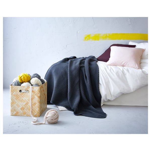 A cosy bedroom scene featuring a blue woven cotton VÅRELD bedspread, pink and purple pillows, and a basket with colourful yarn.