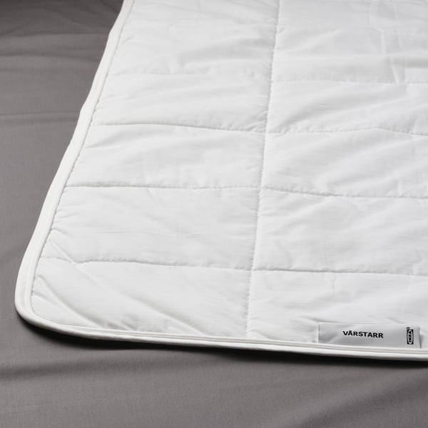 A white duvet with square quilting lies on a grey bed. It a subtle jacquard stripe design and a tag labelled VÅRSTARR and IKEA at the corner.