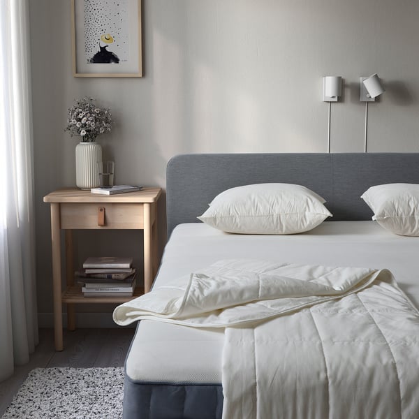 Serene bedroom with twin bed, white duvet, blue headboard, bedside table, books, flowers, wall lights, and rug.