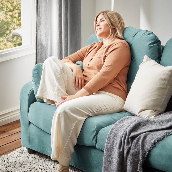 A person relaxes on a teal VINLIDEN sofa, covered with a beige blanket, near a window with sheer curtains.