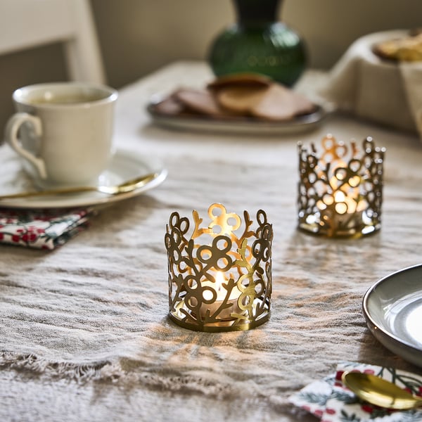 Golden VINTERFINT tea light holder, stainless steel with acrylic coating, intricate mistletoe design, small size, on tablecloth among other items.